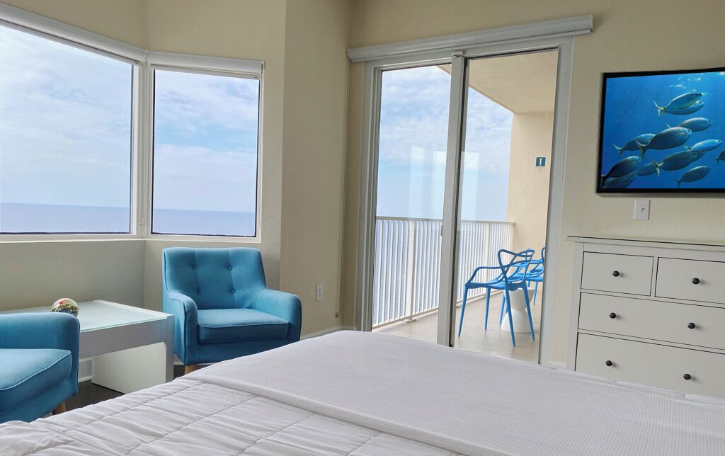 Habitación de hotel con cama, escritorio, silla, televisor y balcón con vista al mar.