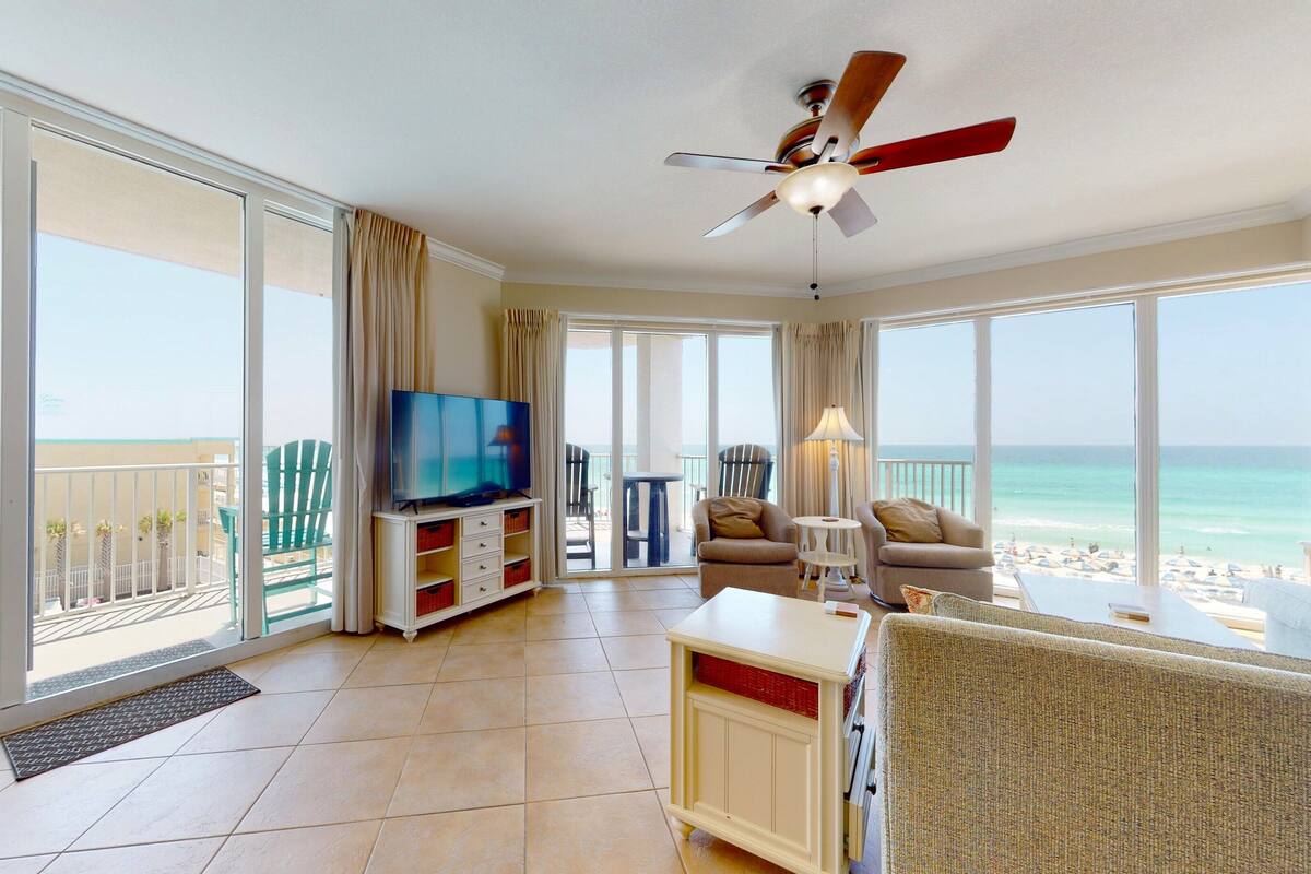 Ein großzügiges Wohnzimmer mit großem Fernseher, bequemen Sitzmöglichkeiten und einem Balkon mit Blick auf Strand und Meer.