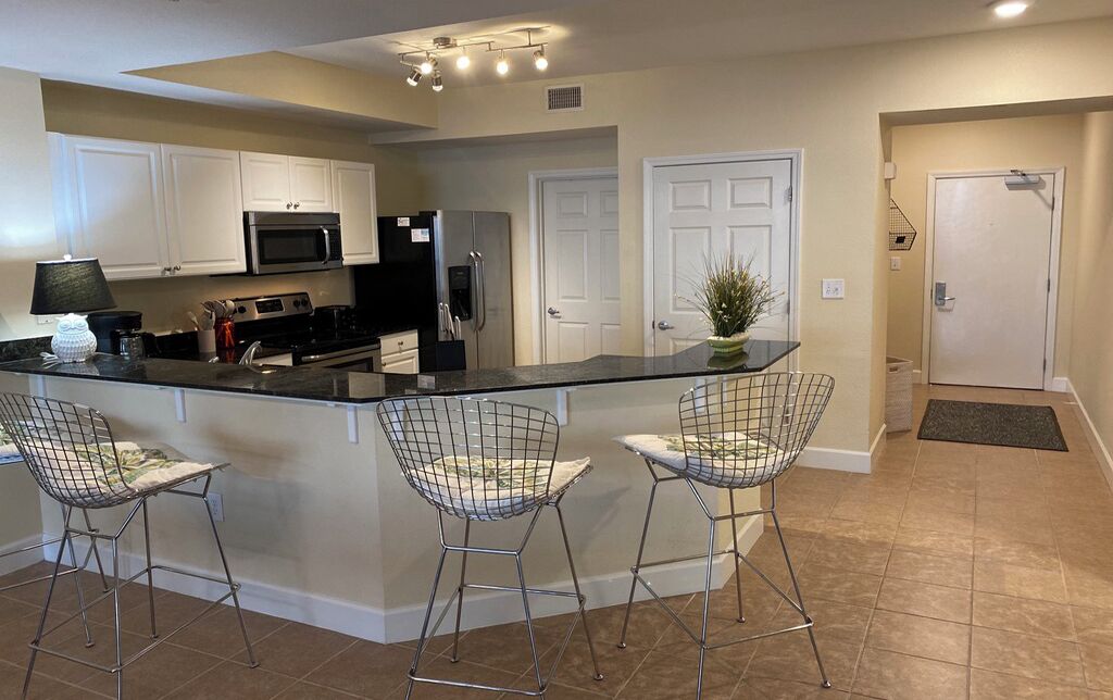 A modern kitchen with white cabinets, stainless steel appliances, a black countertop, and three wire-framed bar stools.