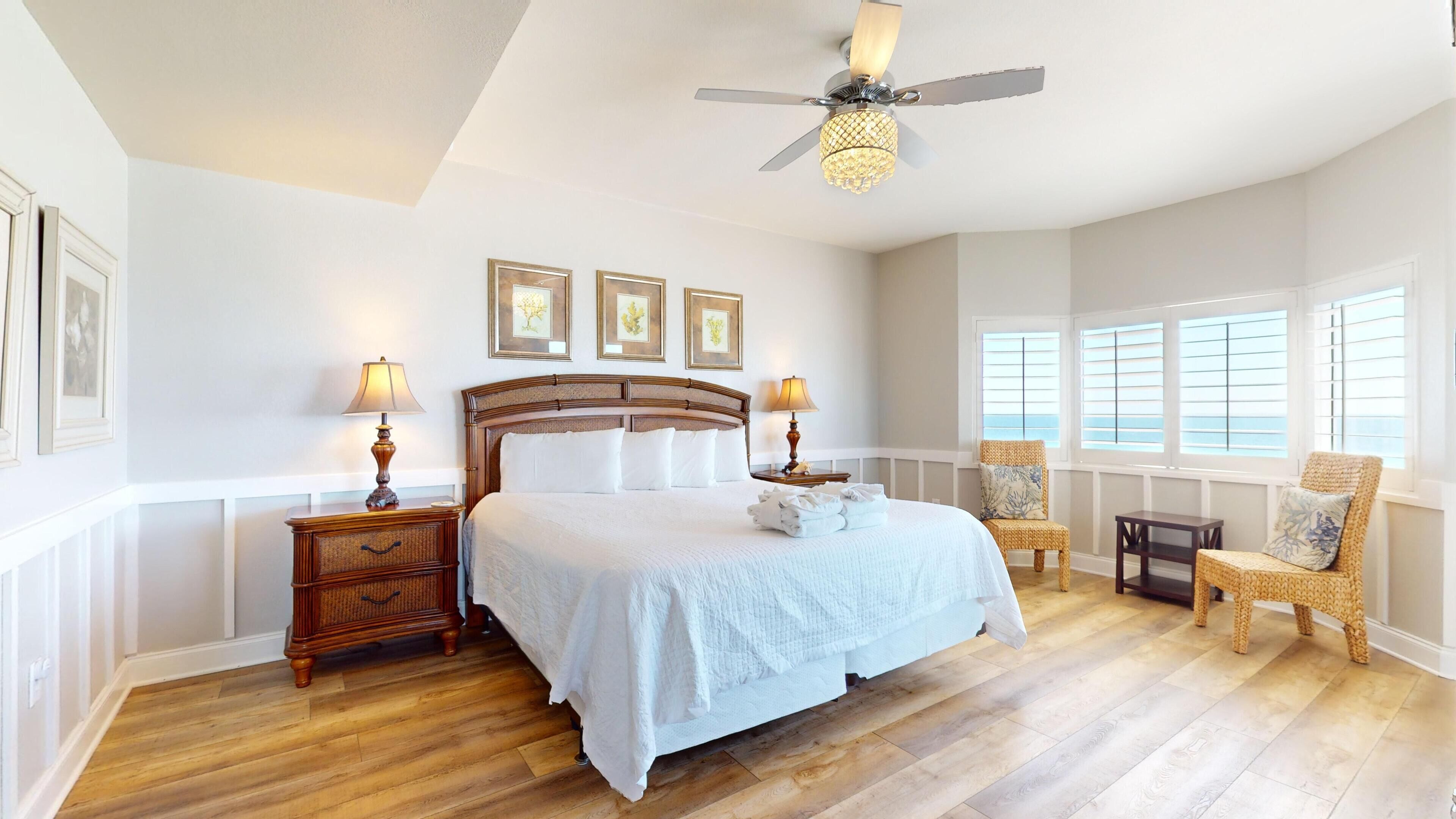 A spacious bedroom with a large bed, two nightstands, a ceiling fan, and a view of the ocean through the windows.
