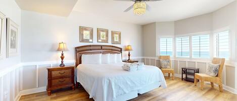 A spacious bedroom with a large bed, two nightstands, a ceiling fan, and a view of the ocean through the windows.