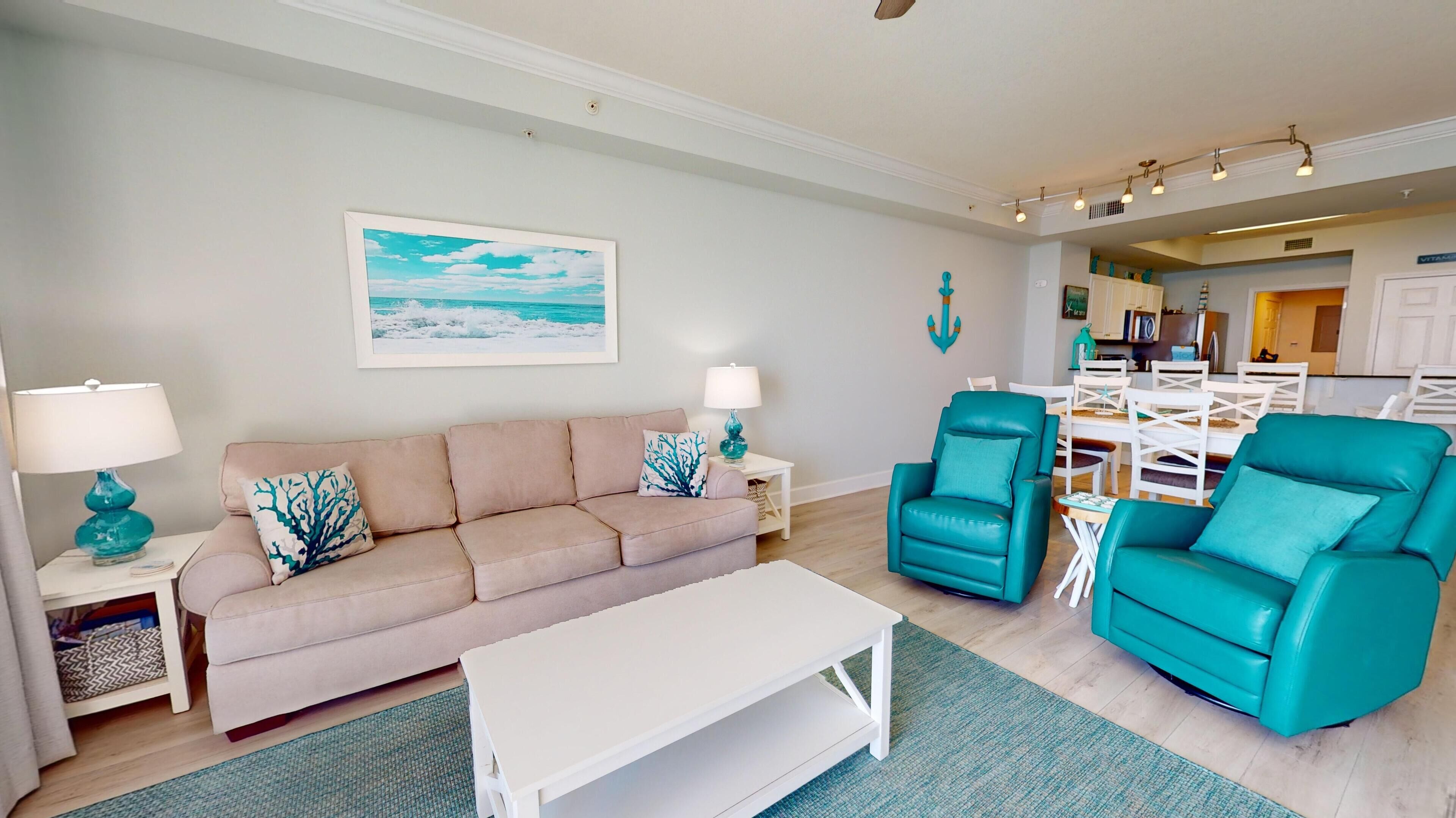 A living room with a beige sofa, teal armchairs, a white coffee table, and a painting of a beach scene.