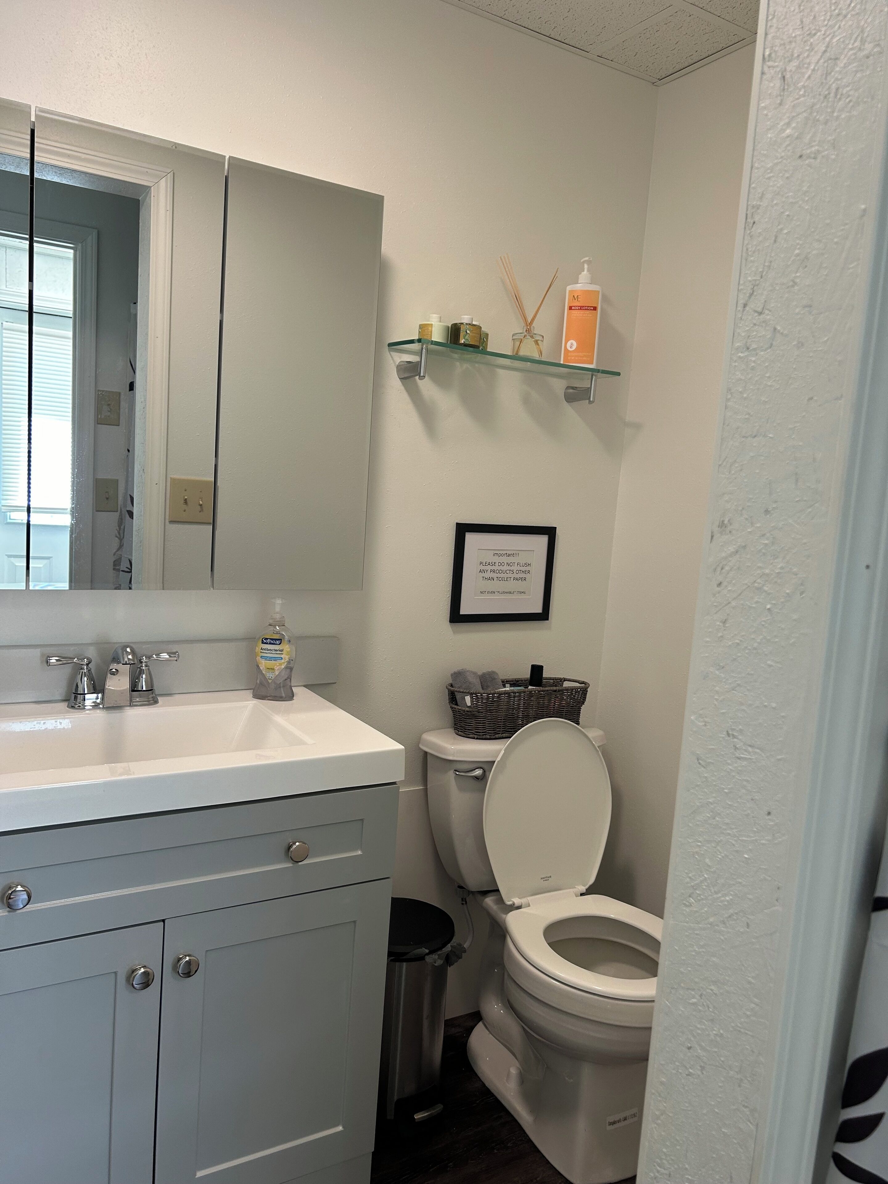 Ein Badezimmer mit einem weißen Waschbecken, Spiegel und WC. An der Wand hängt ein gerahmtes Schild und ein Regal mit Toilettenartikeln.