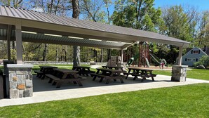 Un espace pique-nique abrité, doté de plusieurs bancs et de tables à pique-nique, entouré d’une pelouse verdoyante et d’arbres.