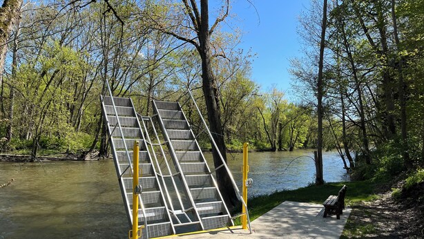 Un chemin piétonnier en béton, bordé de rampes m étalliques, mène à une rivière, le tout étant entouré d’arbres et agrémenté d’un banc.