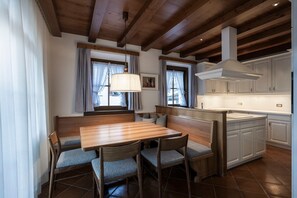 A dining area with a wooden table and chairs, a bench, and a kitchen with white cabinets and a range hood. - Apartment 'Appartamento Stube' with Mountain View, Private Terrace and Wi-Fi (Ortisei)