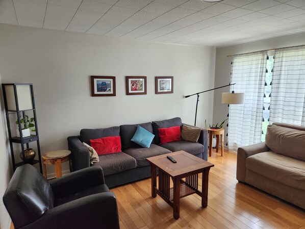 Ett vardagsrum med en mörkgrå soffa, ett soffbord i trä och en beige fåtölj. På väggen hänger tre inramade tavlor, och det finns en golvlampa och en hög växt i en hylla.