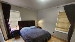 Un dormitorio con piso de madera, una cama oscura con cabecera, dos mesitas de noche y un buró.