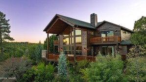 Une maison en bois à deux étages, dotée d’un balcon et de grandes fenêtres, entourée d’une végétation luxuriante.