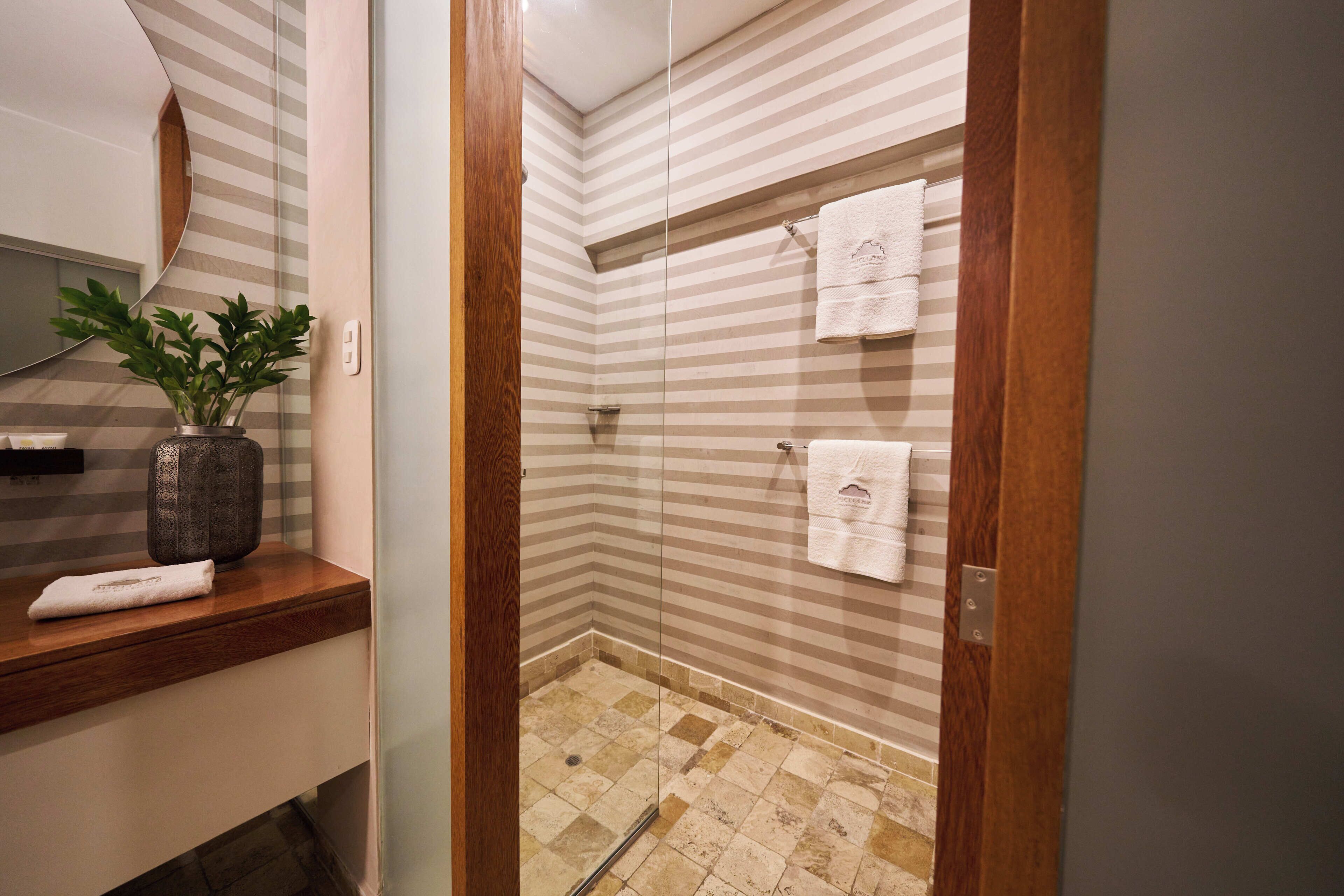 A modern bathroom with a glass shower enclosure, striped wall tiles, and a wooden vanity with a plant and towels.