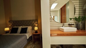 Habitación de hotel con cama y tablero de cabecera capitonado, lámparas de noche, un espejo grande y una tocador de madera con lavabo.