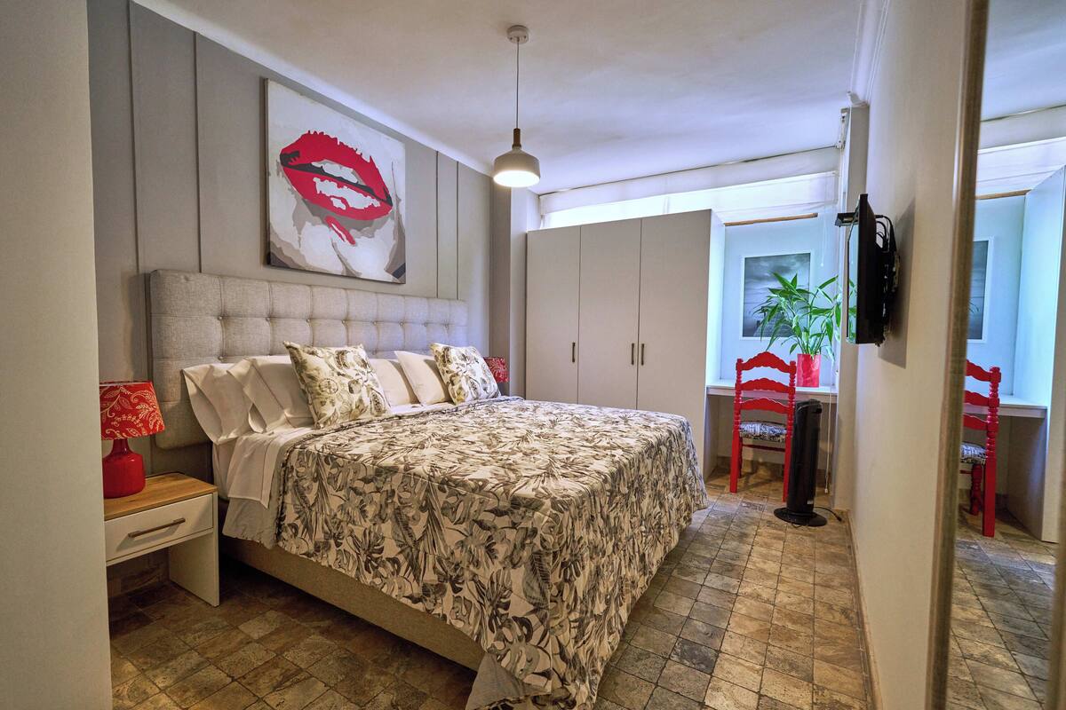 A bedroom with a bed, a nightstand, a red lamp, a large floral bedspread, a wall-mounted TV, and a window with a plant.