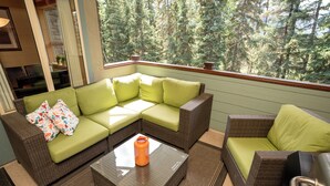 A cozy outdoor seating area with a green sofa and a wicker chair, a glass coffee table, and a view of a forest. - Relax with a Cozy Private Deck, Warm South Sun and Pool & Hot Tubs. (Canmore)
