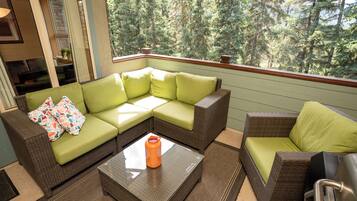 A cozy outdoor seating area with a green sofa and a wicker chair, a glass coffee table, and a view of a forest.
