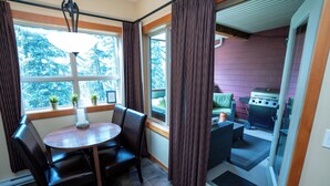A dining area with a round table and chairs, a window with curtains, and a view of the outdoors. - Relax with a Cozy Private Deck, Warm South Sun and Pool & Hot Tubs. (Canmore)