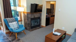 A living room with a blue armchair, a fireplace, a television mounted above it, and a small wooden coffee table. - Relax with a Cozy Private Deck, Warm South Sun and Pool & Hot Tubs. (Canmore)