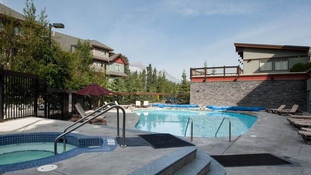 A swimming pool with lounge chairs, a red umbrella, and a stone wall. - Relax with a Cozy Private Deck, Warm South Sun and Pool & Hot Tubs. (Canmore)