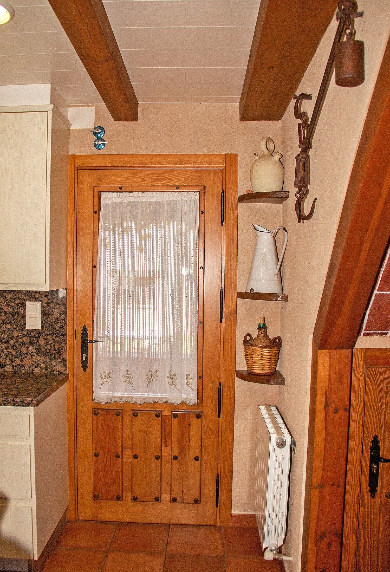 Une porte en bois avec un rideau blanc, un radiateur, des étagères remplies d’ustensiles de cuisine et un luminaire suspendu.