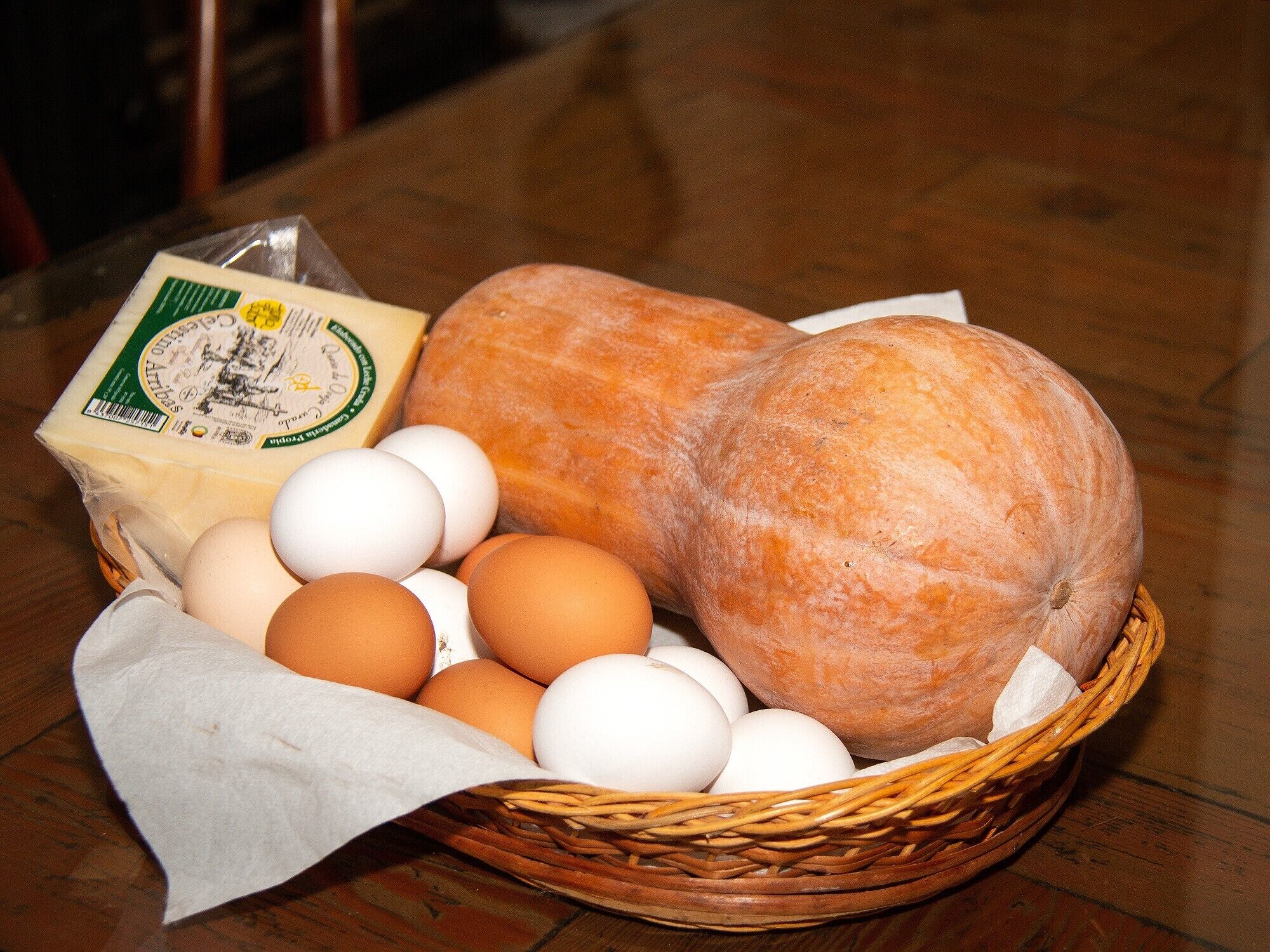 Una cesta con huevos, una hogaza de pan y un bloque de queso sobre una mesa de madera.