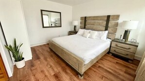 Un dormitorio con una cama de madera, una mesita de noche con lámpara, un espejo y una planta en maceta.