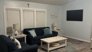 A living room with a dark-colored sofa, a coffee table, and a TV mounted on the wall. - Coastal Cottage - 2BR Home with 2 Pools, pond, & 1000ft pier in Assateague Point (Berlin)