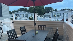 A patio with a table and chairs, an umbrella, and a view of mobile homes in the background. - Coastal Cottage - 2BR Home with 2 Pools, pond, & 1000ft pier in Assateague Point (Berlin)