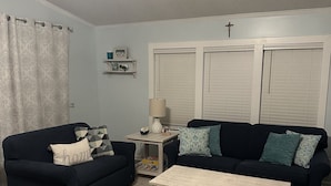 A living room with two dark sofas, a small table, and a coffee table. There is a window with blinds, a curtain, and a cross on the wall. - Coastal Cottage - 2BR Home with 2 Pools, pond, & 1000ft pier in Assateague Point (Berlin)