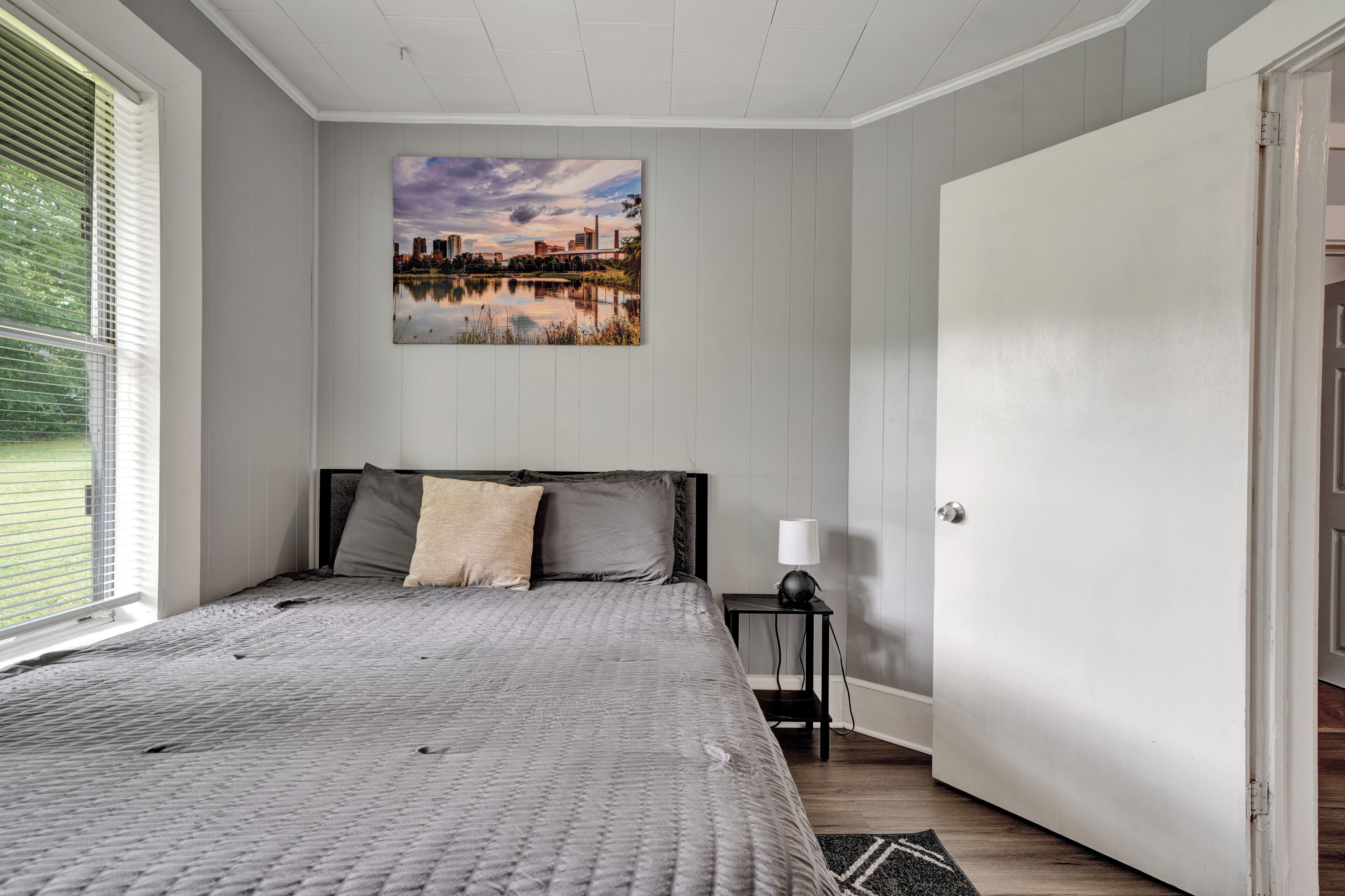 A bedroom with a bed, a nightstand, a lamp, a door, and a cityscape painting on the wall.