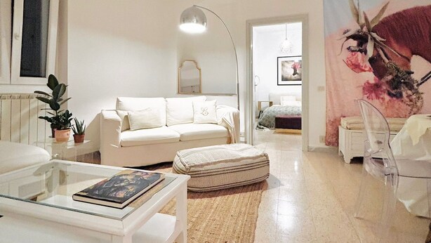 Una sala de estar moderna con un sofá blanco, una mesa de centro de vidrio y una silla transparente. Hay un cuadro grande en la pared y una lámpara de pie.