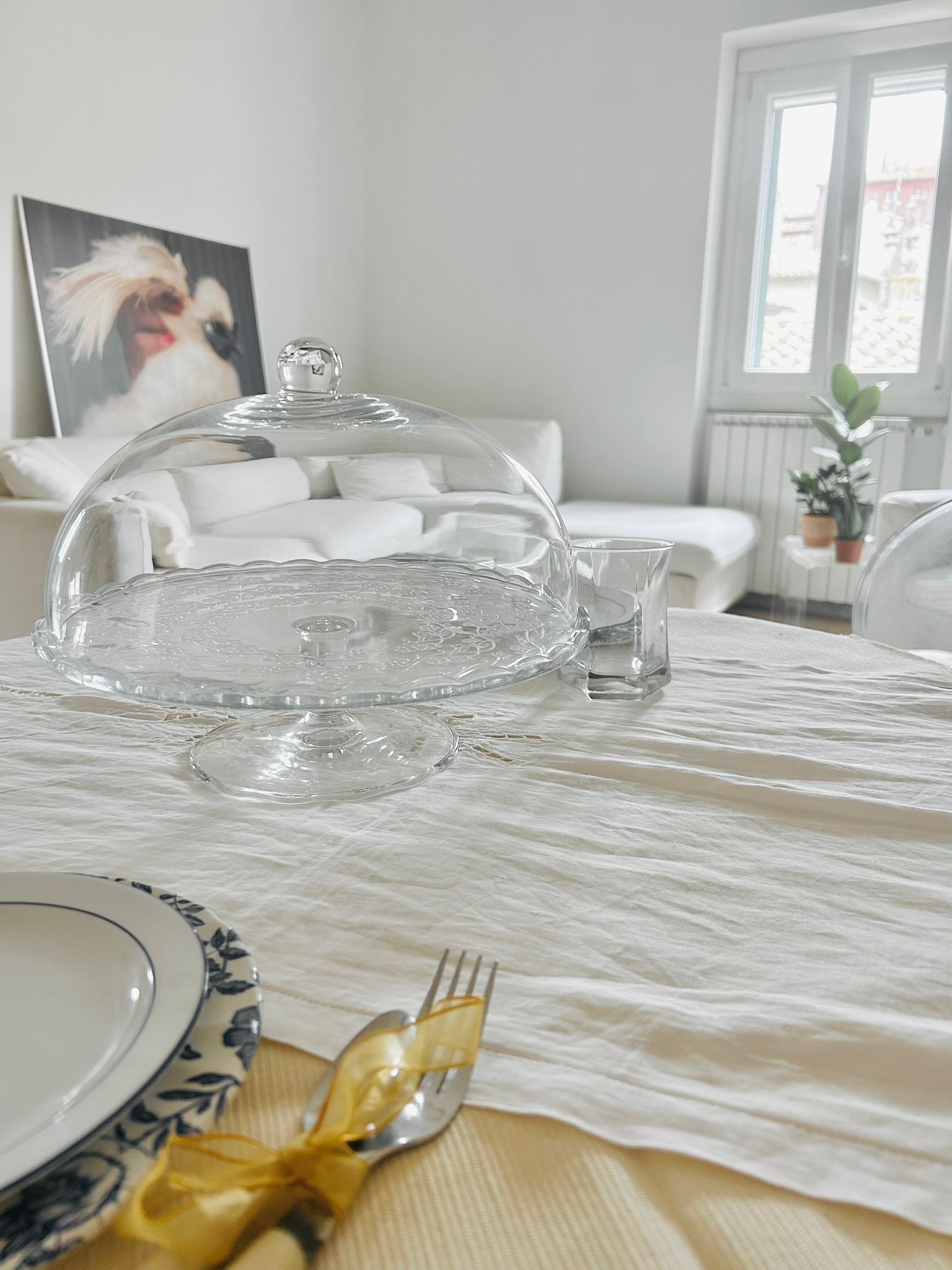 Ein Esstisch mit einer transparenten Glaskuppen-Mitte und einer weißen Tischdecke, dazu ein kleines Glas Wasser.