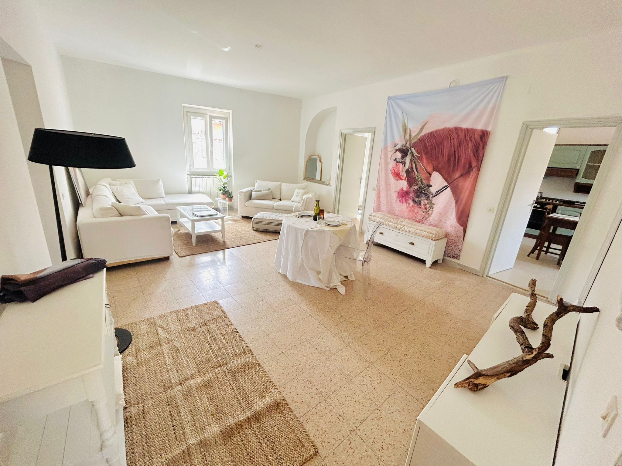 A spacious living room with a white sofa, a coffee table, and a dining table set for four. A large floral tapestry hangs on the wall, and a kitchen is visible through an open doorway.