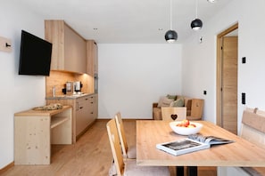 A modern kitchen with wooden cabinets, a dining table with chairs, a TV mounted on the wall, and pendant lights.
