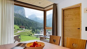 Un coin repas avec une table en bois, un saladier de fruits et une vue sur les montagnes et un village, le tout visible à travers de grandes fenêtres.