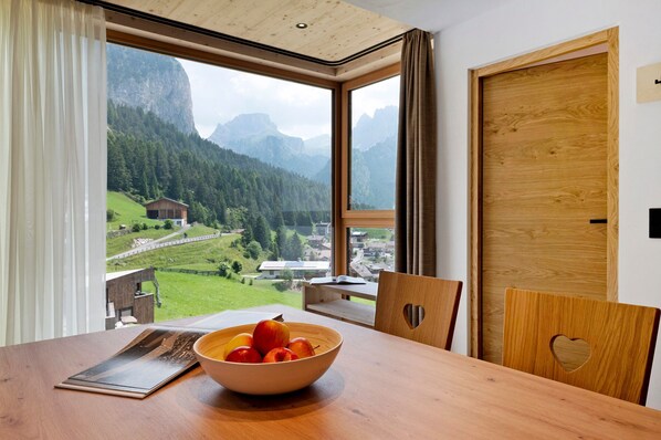 A dining area with a wooden table, a bowl of fruit, and a view of mountains and a village through large windows.