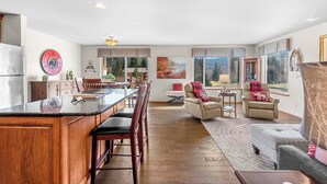 A spacious living area with a large wooden dining table, multiple chairs, a sofa, and a coffee table. There is a kitchen area with a refrigerator and cabinets. The room has large windows with curtains, and a fireplace is visible in the background. - Golf Course View Condo • Quick Walk to Downtown Leavenworth & Resort Pools! (Leavenworth)
