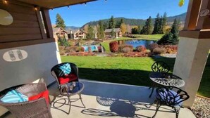An outdoor patio with wicker chairs and a small round table, overlooking a scenic landscape with a lake and houses. - Golf Course View Condo • Quick Walk to Downtown Leavenworth & Resort Pools! (Leavenworth)