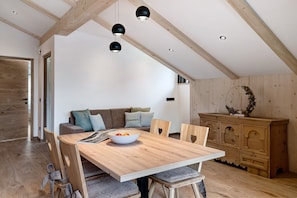 A dining area with a wooden table, chairs, and a bowl of fruit. There is a sofa with cushions and a wooden cabinet in the background.
