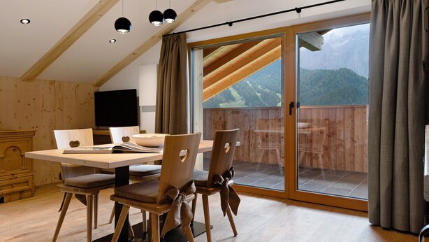 A dining area with a wooden table and chairs, a television, and a view of a mountainous landscape through a sliding glass door. - Apartment 'Fën' with Mountain View, Shared Garden and Wi-Fi (Wolkenstein)