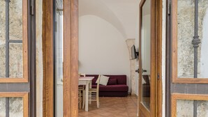 Um quarto com porta de madeira, uma mesa de jantar com cadeiras, um sofá vermelho e uma TV fixada na parede.