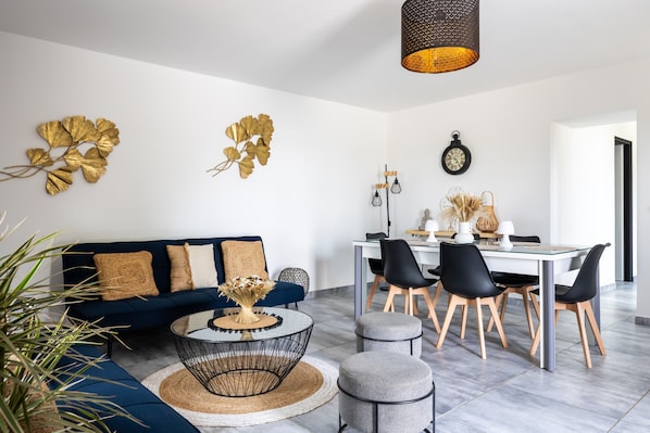 Un salon moderne avec un canapé bleu foncé, une table basse ronde et un coin repas comprenant une table blanche et des chaises noires.
