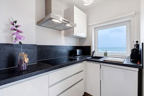 A modern kitchen with a black countertop, white cabinets, a built-in oven, and a window with a view of the sea.