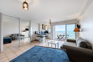 A modern hotel room with a bed, a sofa, a dining area, and a view of the sea.