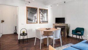 A modern living room with a round wooden table, white chairs, a blue sofa, and a green armchair. There are two framed black and white photographs on the wall, a flat-screen TV, and a fireplace.