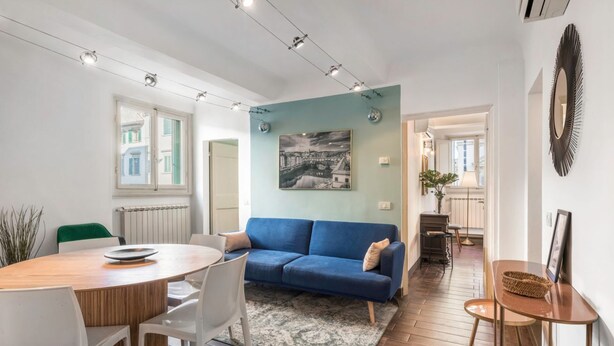 A modern living room with a blue sofa, a round wooden dining table, and a large window.