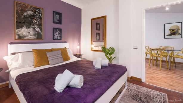 A modern bedroom with a large bed, a bedside table, a mirror, and a dining area with yellow chairs. - Heart of Florence: Central, Cozy Apt with Elevator (Firenze)