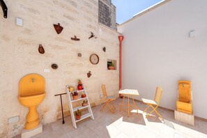 Un petit patio extérieur avec un mur blanc, un évier jaune, une échelle blanche, une chaise en bois, une table et une petite étagère avec des plantes.