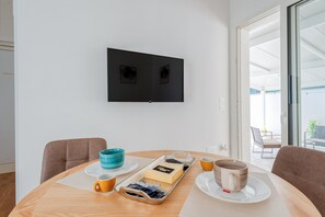A modern room with a flat-screen TV mounted on the wall, a round dining table with a tray of breakfast items, and a view of the outdoors through a glass door.