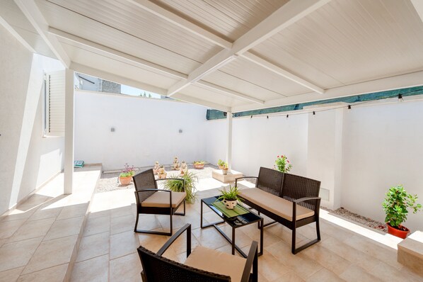 Une terrasse extérieure dotée d’un espace salon couvert, de plantes en pot et d’un sol carrelé.