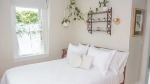 Un lit bien fait, avec du linge de lit blanc et des oreillers, une fenêtre ornée de rideaux à motifs floraux et une étagère murale décorée de plantes et d’un cadre photo.