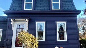 Une maison à deux étages, avec une façade bleu foncé, des encadrements de fenêtres et de portes blancs, et un petit porche doté d’une porte blanche.
